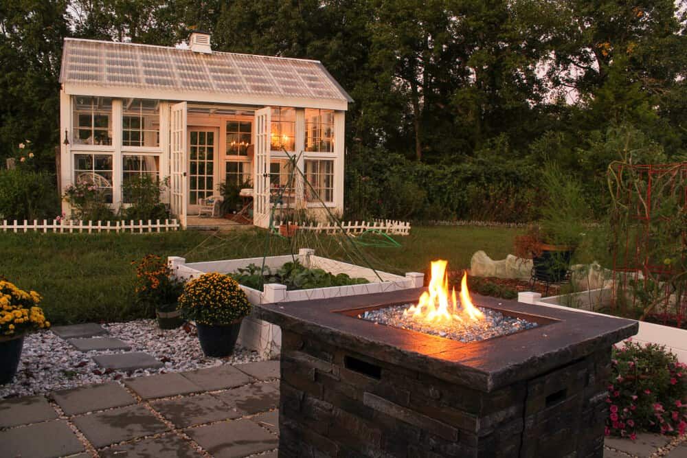 A backyard in the early evening with a fire burning in a square pit and a bright greenhouse behind it, surrounded by plants and flowers